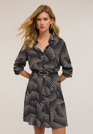 Robe chemise noire et blanche à motifs, avec un devant à boutons, des manches retroussées et une taille cintrée mise en valeur par une ceinture noire et une boucle en or.