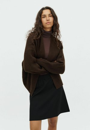 Brown ribbed cardigan with open front, worn over a fitted dark purple turtleneck and black skirt. Soft texture and relaxed fit.