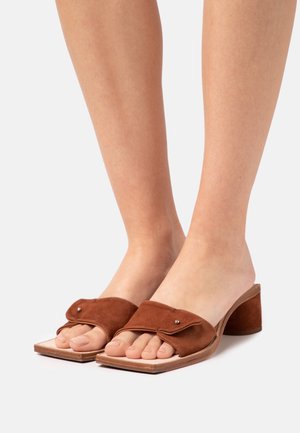 Bruine suede sandalen met hak en open teen, met een brede band en decoratieve hardware. Vierkante vorm en een lage blokhak.