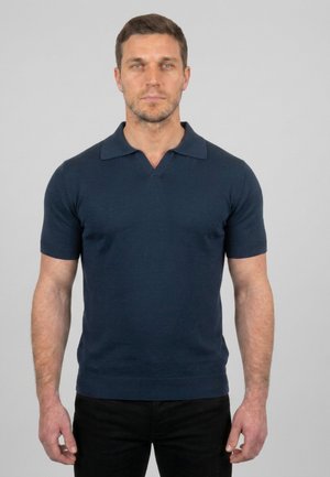 Hombre con cabello corto castaño que lleva una camiseta tipo polo de manga corta azul marino y pantalones negros, parado frente a un fondo gris liso.