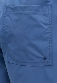 Gros plan d'une poche arrière en tissu bleu avec des coutures renforcées et un petit œillet de ventilation près du coin inférieur droit.