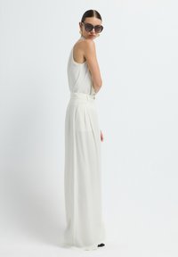 Witte, mouwloze top gecombineerd met hoge taille, wijde broek met plooien en een knoopdetail. Model draagt een zonnebril en oorbellen.