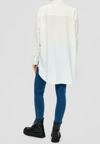 White long-sleeve shirt with a curved hem, buttoned cuffs, and a back yoke. Paired with dark blue skinny jeans and black chunky boots.
