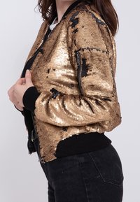Giacca bomber in paillettes dorate con dettagli neri; presenta una chiusura a zip, polsini a costine e una vestibilità leggermente cropped. Superficie testurizzata con luccichio.