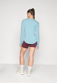 Helles, blaues Langarmoberteil mit rundem Halsausschnitt und abgerundetem Saum, kombiniert mit kastanienbraunen Shorts. Person trägt weiße Socken und helle Sneakers.