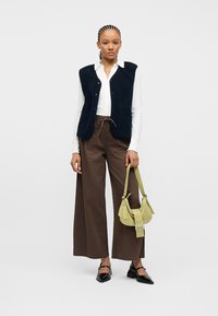 Γυναίκα με πλεξούδες, φορώντας λευκό πουκάμισο, μαύρο γιλέκο, καφέ παντελόνια με φαρδιά μπατζάκια, μαύρα παπούτσια, κρατώντας μια μικρή τσάντα σε ανοιχτό πράσινο χρώμα.