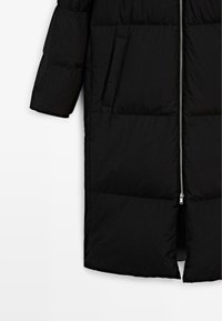 Zwarte pufferjas met ritssluiting, verticale naden en zijzakken. Heeft een knielange snit en een matte textuur.