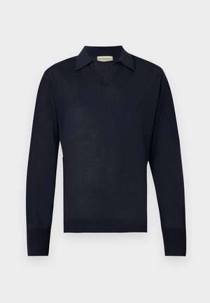 Man staand, draagt een marineblauw overhemd met lange mouwen en kraag, grijze geplooide broek en zwarte nette schoenen, handen in de zakken, kijkt zijwaarts.