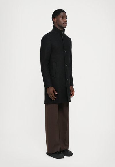 Abrigo de lana negro con cuello alto, botones frontales y bolsillos laterales, llevado sobre pantalones marrones holgados y zapatos negros.
