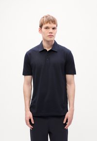 Joven con cabello corto castaño claro, vistiendo una camisa polo negra de manga corta y pantalones negros a juego, de pie contra un fondo blanco.