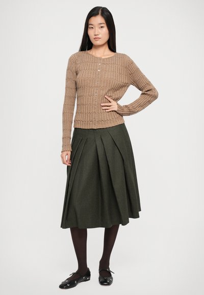 Cardigan maro cu nervuri și nasturi, purtat cu o fustă plisată verde. Modelul poartă pantofi negri și colanți negri, stând în picioare.