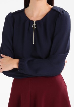 Woman wearing a navy blue long-sleeve blouse with a front zipper and a maroon skirt, arms crossed, against a white background.