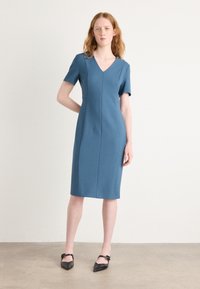 V-neck, short-sleeve dress in textured blue fabric, fitted silhouette, knee-length, featuring vertical seams and model wearing black pointed flats.