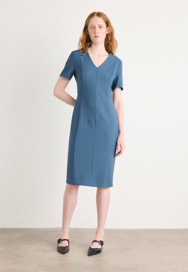 V-neck, short-sleeve dress in textured blue fabric, fitted silhouette, knee-length, featuring vertical seams and model wearing black pointed flats.