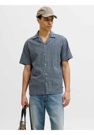 Jeune homme portant une casquette beige, une chemise à manches courtes à carreaux bleus et un jean bleu clair, tenant un sac beige et noir dans sa main gauche.