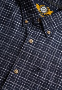 Camisa de cuadros azul marino con botones beige y logo bordado en el bolsillo del pecho.