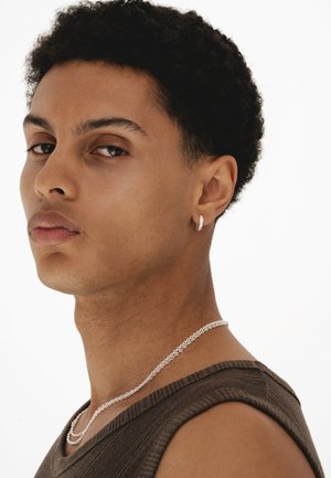 Bărbat tânăr cu păr scurt și creț, purtând un tricou maro fără mâneci, cercei argintii în formă de cerc și un lănțișor argintiu, privind într-o parte.