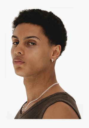 Nuori mies, jolla on lyhyet kiharat hiukset, hänellä on päällään ruskea hihaton paita, hopeinen renkaankorvakoru ja hopeaketjukaulakoru, katse käännetty sivulle.