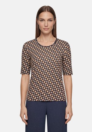 Femme aux cheveux bruns raides porte un haut à manches courtes à motifs avec des formes de feuilles bleu marine et beige et un pantalon bleu foncé, debout devant un fond uni.