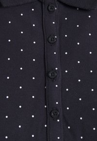 Granatowa koszulka polo z teksturowanej tkaniny, z białymi kropkami i czterema czarnymi guzikami wzdłuż zapięcia z przodu.