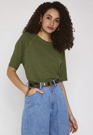 Femme aux cheveux bouclés portant un t-shirt vert à manches courtes, un jean bleu taille haute, une ceinture marron, des boucles d'oreilles anneaux dorés, des bracelets et des bagues sur un fond uni.