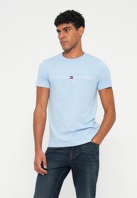 Šviesiai mėlyna medvilninė marškinėliai su trumpomis rankovėmis, apvaliu kaklu, su "TOMMY HILFIGER" logotipu ir balta žemiau esancia įsteigimo metų tekstu.