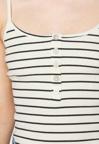 Gestreept tanktop in crèmekleur met zwarte horizontale strepen, voorzien van een knoopsluiting aan de voorkant en dunne bandjes. Getextureerde ribstop.