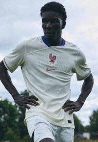 Maglietta da calcio bianca con colletto blu, caratterizzata da un emblema di gallo rosso e dal logo Nike. Maniche corte con strisce colorate ai polsini.