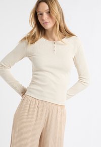 Beżowa bluzka z długim rękawem o prążkowanej fakturze i okrągłym dekoltem, z trzema guzikami przy szyi oraz wzorzyste, luźne spodnie.