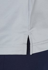 Chemise gris clair avec une texture douce, fentes latérales et détails cousus. Conçue avec un ourlet arrondi, associée à un pantalon bleu marine.