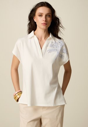 Donna che indossa una camicetta bianca a maniche corte con motivo di palma in paillettes argento sulla spalla, pantaloni beige e braccialetti impilati al polso.