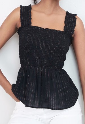 Kobieta w czarnej, bezrękawnikowej bluzce z falbaniastymi ramiączkami i pionowymi metalicznymi paskami, w białych spodniach, z rękami w kieszeniach.