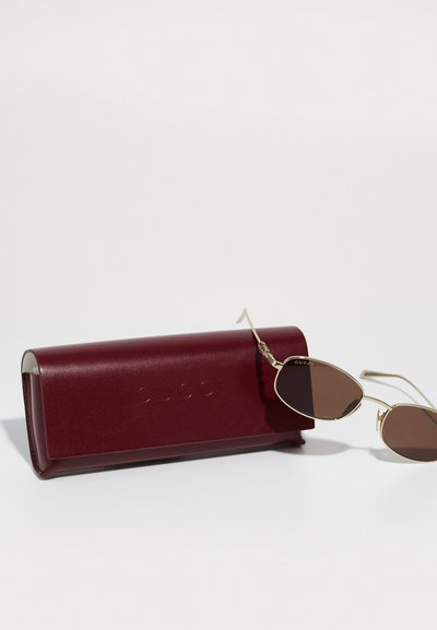 Gafas de sol marrones en forma ovalada con montura dorada, al lado de un estuche rígido burdeos embellecido con "GUCCI". Texturas suaves y detalles de diseño minimalistas.