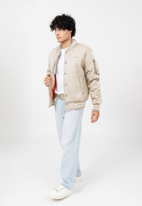 Beżowa bomberka z bocznymi kieszeniami, ściągaczami na mankietach i szarymi paskami. Wyścielona intensywnym pomarańczem. Połączona z jasnoniebieskimi dżinsami i białymi trampkami.