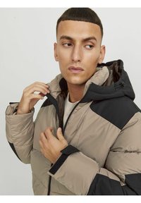 Giacca a vento in beige e nero, con apertura zip, cappuccio e polsini in velcro. La texture è liscia, con un design trapuntato per l'isolamento.