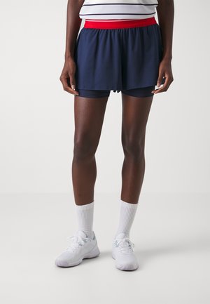 Person trägt marineblaue Sportshorts mit rotem Bund, weiße Sportsocken, weiße Sportschuhe und ein gestreiftes Oberteil, steht vor einem weißen Hintergrund.