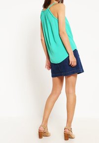 Una blusa sin mangas de color turquesa con detalle fruncido en la parte posterior, combinada con una falda de mezclilla y tacones beige. El conjunto tiene un ajuste relajado.
