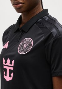 Άτομο που φοράει μαύρη φανέλα του Club Internacional de Fútbol Miami με ροζ λογότυπα και μαύρο γιακά, εμφανίζοντας το πάνω μέρος του σώματος και τα χείλη.