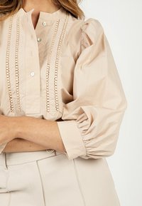 Blouse beige clair à manches ballon, fermeture par boutons, avec détails décoratifs en découpes le long du plastron avant. Matière en coton doux.