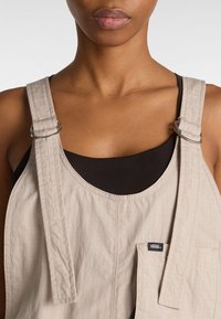Beige Latzhose mit Trägern und silbernem Metall, einer Brusttasche und einem strukturierten Stoff. Unterhalb ist ein schwarzes Tanktop sichtbar.