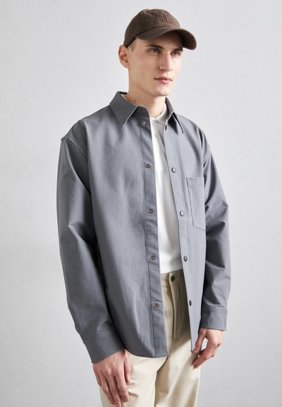 Gray button-up shirt in a relaxed fit, with a chest pocket and silver hardware. Worn over a white polo, paired with beige pants.