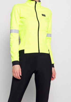 Veste de sport jaune vif avec un col haut, fermeture éclair et accents réfléchissants. Associée à des leggings noirs, au design ajusté.