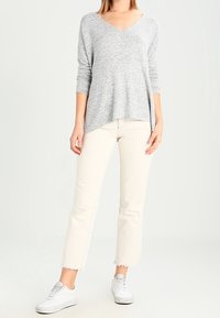 Grauer, langärmeliger Pullover mit V-Ausschnitt und lockerer Passform, kombiniert mit cremefarbenen, cropped Jeans mit fransigen Säumen; weiße Sneakers.