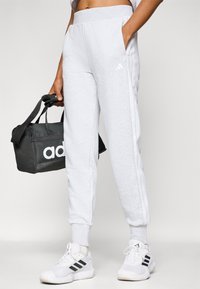 Los pantalones de chándal gris claro tienen una cinturilla ajustada, bolsillos laterales y detalles de rayas blancas. Se combinan con una bolsa negra y zapatillas blancas.