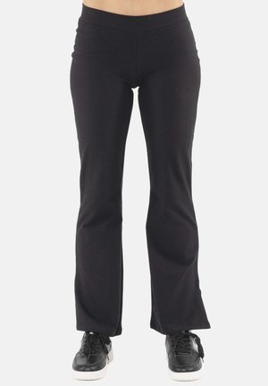 Femme portant un pantalon bootcut noir ajusté et des baskets noires, debout sur un fond blanc uni.