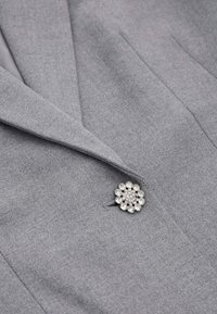Blazer grigio dal tessuto liscio e con rever affilati, accentuato da una spilla a forma di fiore in argento con rhinestones trasparenti.