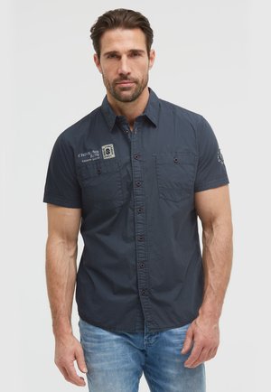Mann trägt ein dunkelblaues kurzärmeliges Hemd mit Knöpfen vorne und Taschen sowie hellblaue Jeans und steht vor einem einfarbigen Hintergrund.