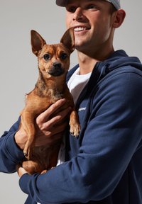 A man in a navy zip-up hoodie holds a small brown dog with large ears. The dog's fur is short and textured with a dark nose.