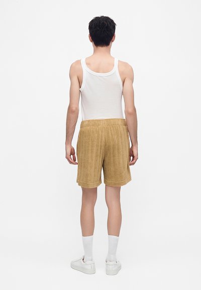 Homme debout de dos portant un débardeur blanc, un short beige texturé, des chaussettes blanches et des baskets blanches sur fond blanc.