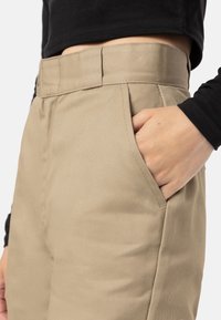 Pantalon beige taille haute avec plis et poches latérales, fabriqué en tissu lisse. La ceinture est structurée, et un haut noir est partiellement visible.